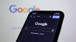 A smartphone displays the Google search page in dark mode, with the Google logo shown on a computer screen in the background.