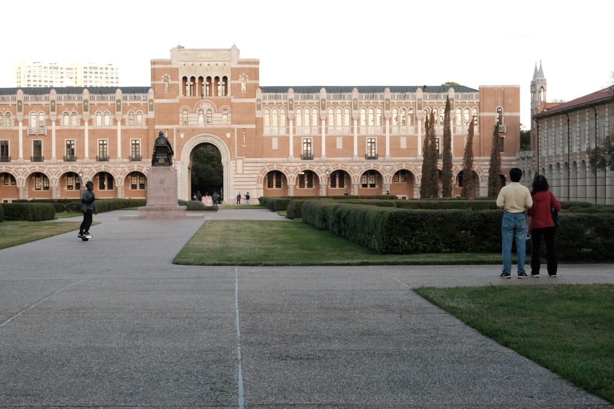 People walk and stand near hedges in front of a large, arched brick university building with a statue in the courtyard.