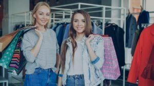 Two women stand in a clothing store holding shopping bags and smiling at the camera. Clothing racks are visible in the background.