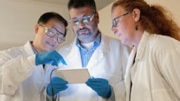 Three scientists in lab coats and gloves examine data on a tablet together in a laboratory setting.