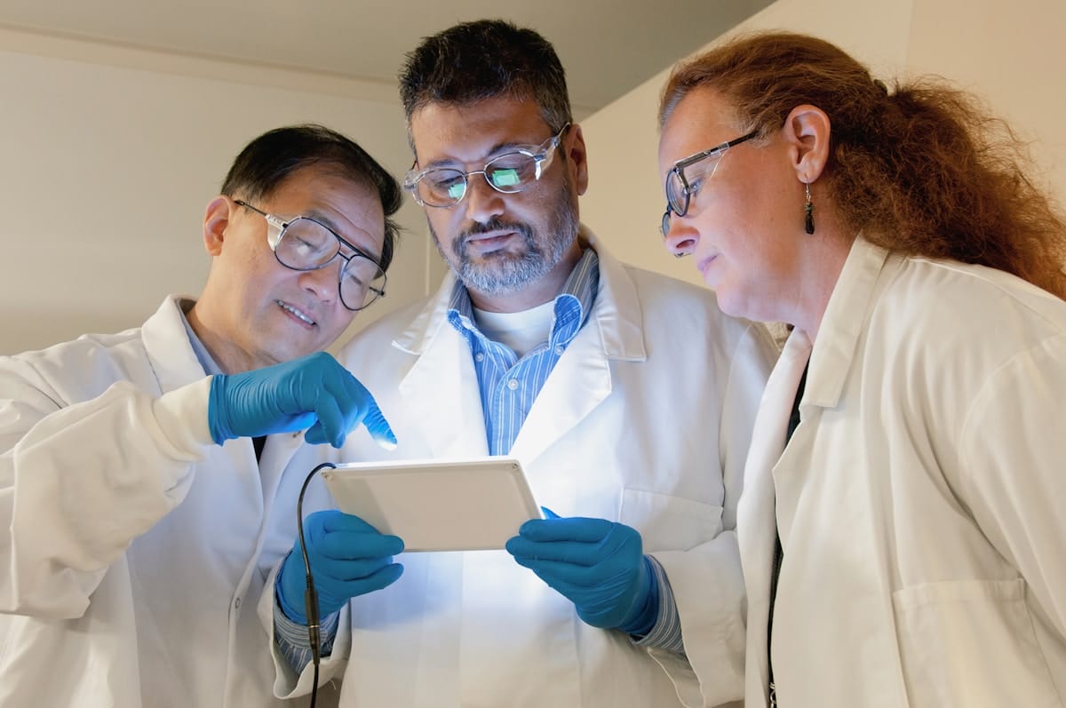 Three scientists in lab coats and gloves examine data on a tablet together in a laboratory setting.