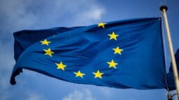 The European Union flag with twelve yellow stars in a circle on a blue background, waving against a partly cloudy sky.