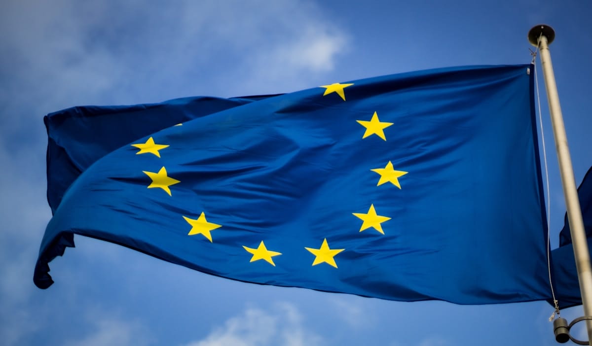 The European Union flag with twelve yellow stars in a circle on a blue background, waving against a partly cloudy sky.
