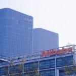 Modern office buildings with the Alibaba logo in the foreground and two tall glass towers in the background under a hazy sky.