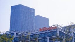 Modern office buildings with the Alibaba logo in the foreground and two tall glass towers in the background under a hazy sky.