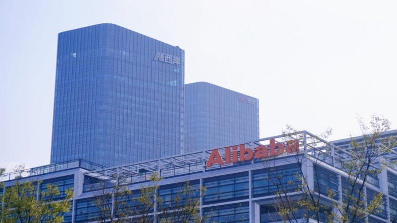Modern office buildings with the Alibaba logo in the foreground and two tall glass towers in the background under a hazy sky.