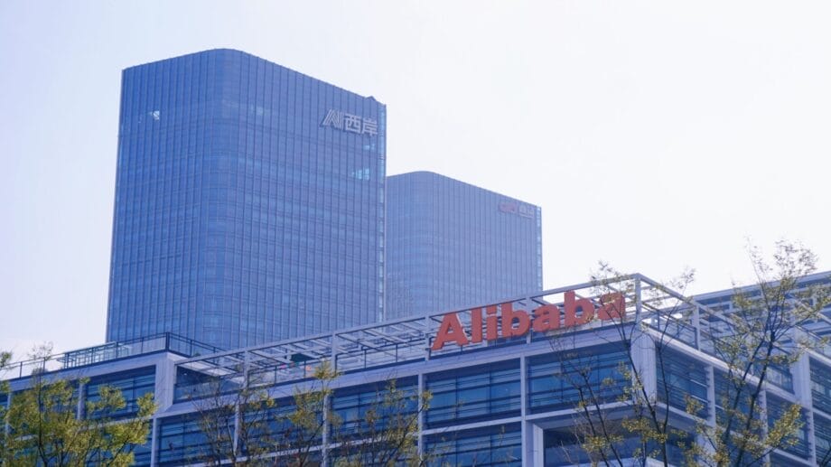 Modern office buildings with the Alibaba logo in the foreground and two tall glass towers in the background under a hazy sky.