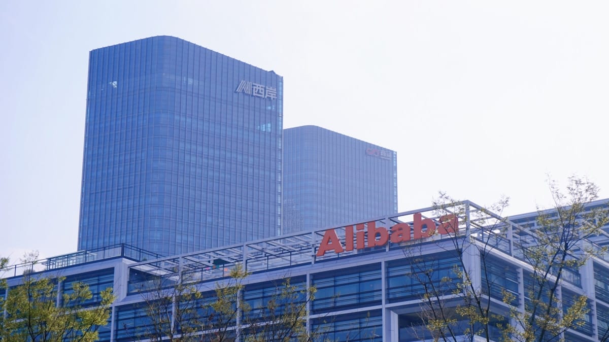 Modern office buildings with the Alibaba logo in the foreground and two tall glass towers in the background under a hazy sky.