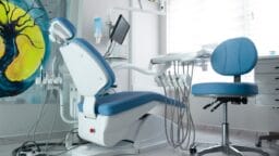 A modern dental office with a blue dental chair, equipment, and a stool in a bright, clean room with a large abstract painting on the wall.