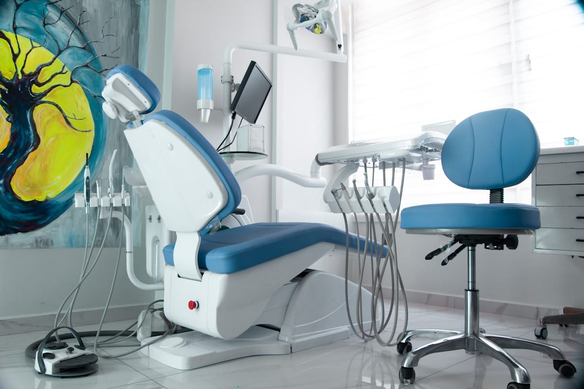 A modern dental office with a blue dental chair, equipment, and a stool in a bright, clean room with a large abstract painting on the wall.