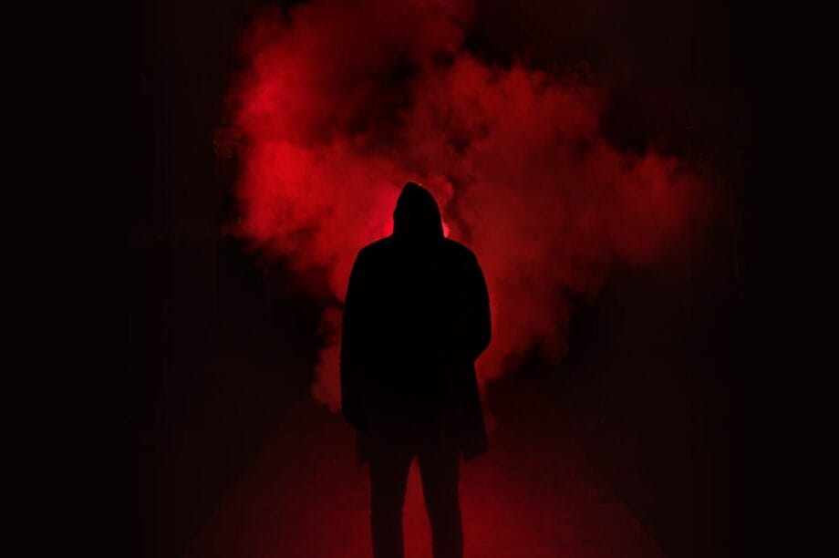 Silhouette of a person in a hooded jacket standing in front of bright red smoke in a dark environment.