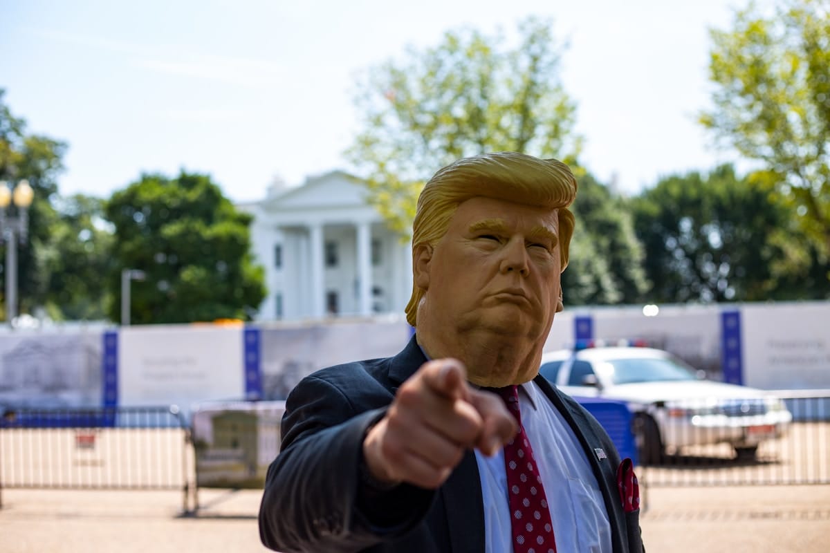 A person wearing a Donald Trump mask and suit stands in front of the White House, pointing toward the camera.