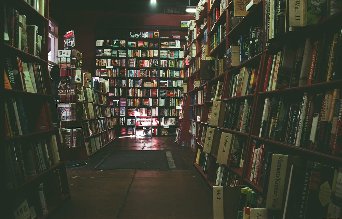 A dimly lit bookstore aisle with shelves filled with books on both sides and more books visible in the background.