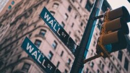 Two street signs for Wall Street and a traffic light are pictured with a tall building blurred in the background.