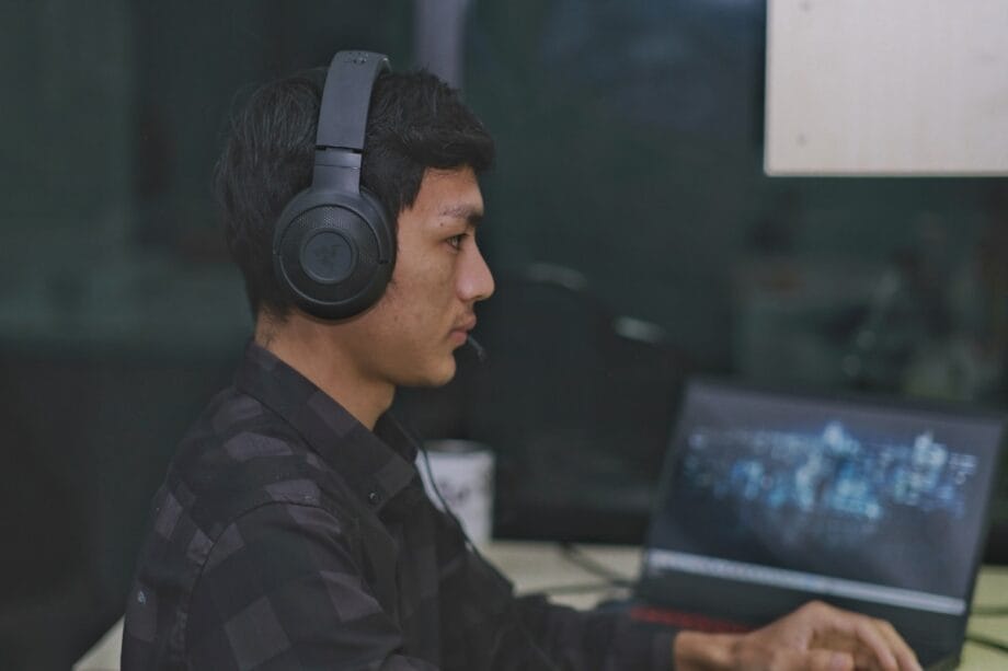 Person wearing headphones and a plaid shirt sits at a desk using a laptop with a blurred cityscape on the screen.