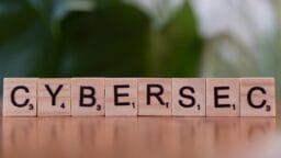 Wooden Scrabble tiles on a table spell out CYBERSEC with a blurred green background.