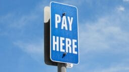 A blue sign with white text reading PAY HERE is mounted on a metal pole against a blue sky.