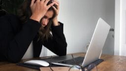 A person sits at a desk, holding their head in frustration while looking at a laptop, with glasses and a phone on the table.