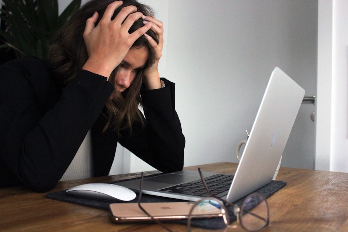 A person sits at a desk, holding their head in frustration while looking at a laptop, with glasses and a phone on the table.