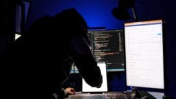 A person in a dark room works at a computer with multiple monitors displaying code and technical information.