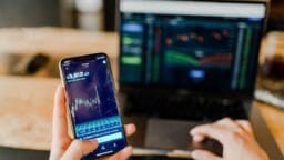 Person holding smartphone with a financial graph, in front of a laptop displaying stock market charts.