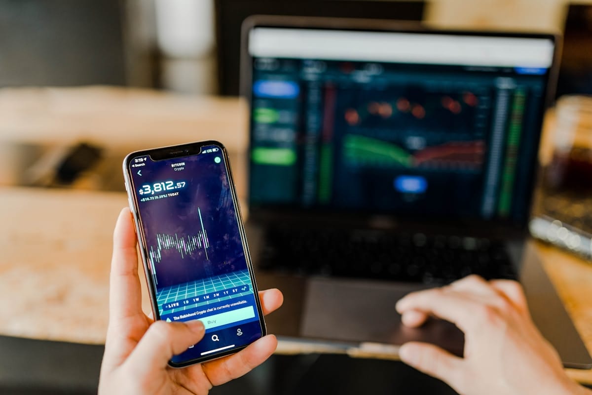 Person holding smartphone with a financial graph, in front of a laptop displaying stock market charts.