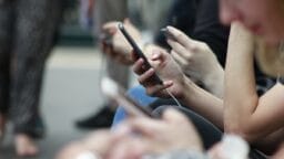 Several people sit in a row, holding and using smartphones, with the focus on their hands and devices.