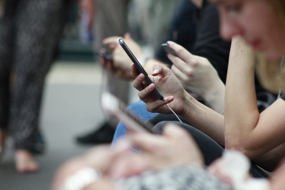 Several people sit in a row, holding and using smartphones, with the focus on their hands and devices.