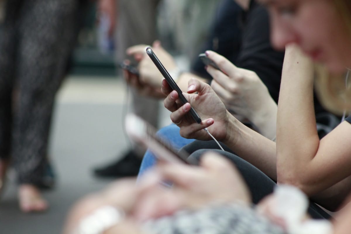 Several people sit in a row, holding and using smartphones, with the focus on their hands and devices.