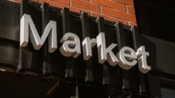 A sign reading Market is mounted on a black fixture attached to a brick wall.