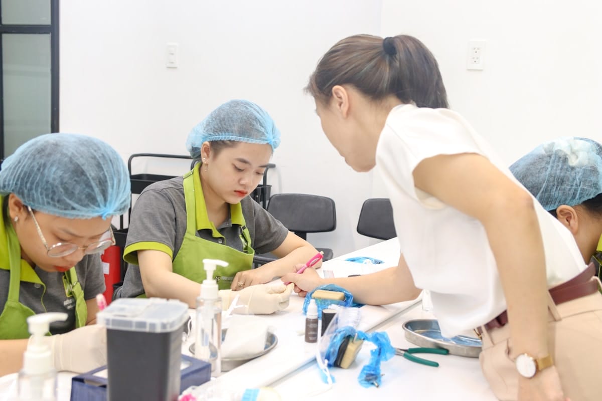 A woman instructs two people wearing green aprons and hairnets as they work on manicure procedures at a table with supplies.