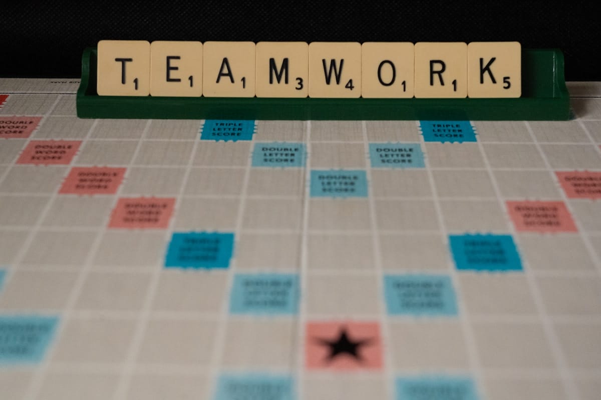 Scrabble tiles spell TEAMWORK on a letter rack above a Scrabble game board with various colored score spaces.