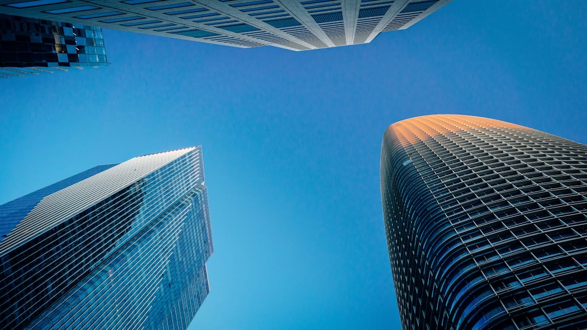 Looking up at three modern skyscrapers against a clear blue sky, with sunlight reflecting off one building.