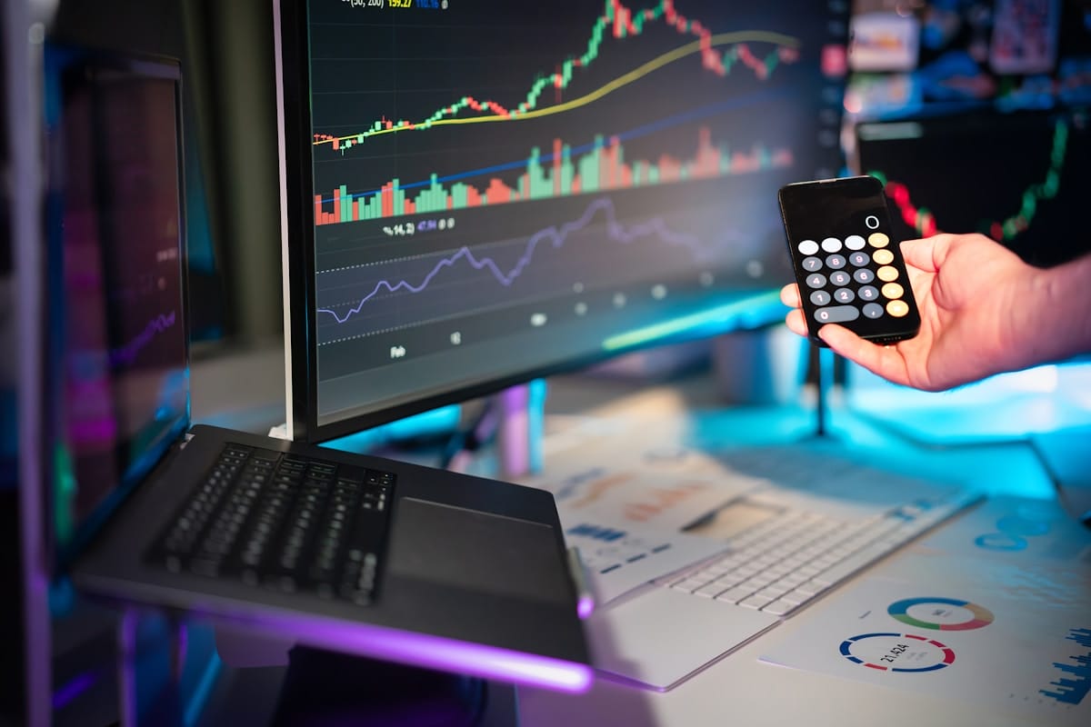 A person holds a calculator in front of computer screens displaying financial charts and a laptop on a desk with papers.