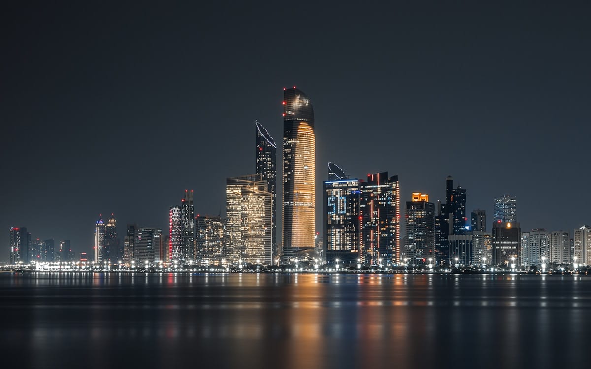 Nighttime cityscape with illuminated skyscrapers reflected on calm water under a clear sky.