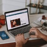 A person uses a laptop at a desk with a camera, notebook, and phone displaying Online Marketing nearby.