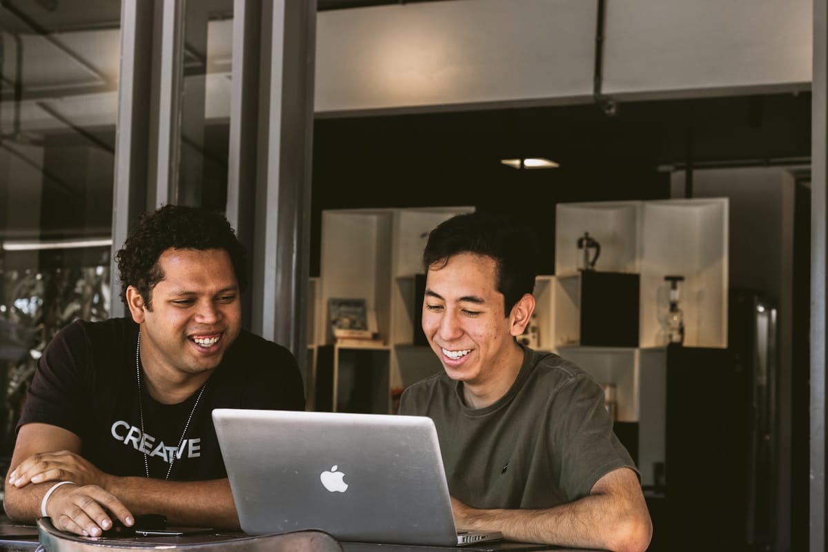 Two people sit at a table, smiling while looking at a laptop screen in a modern indoor setting.