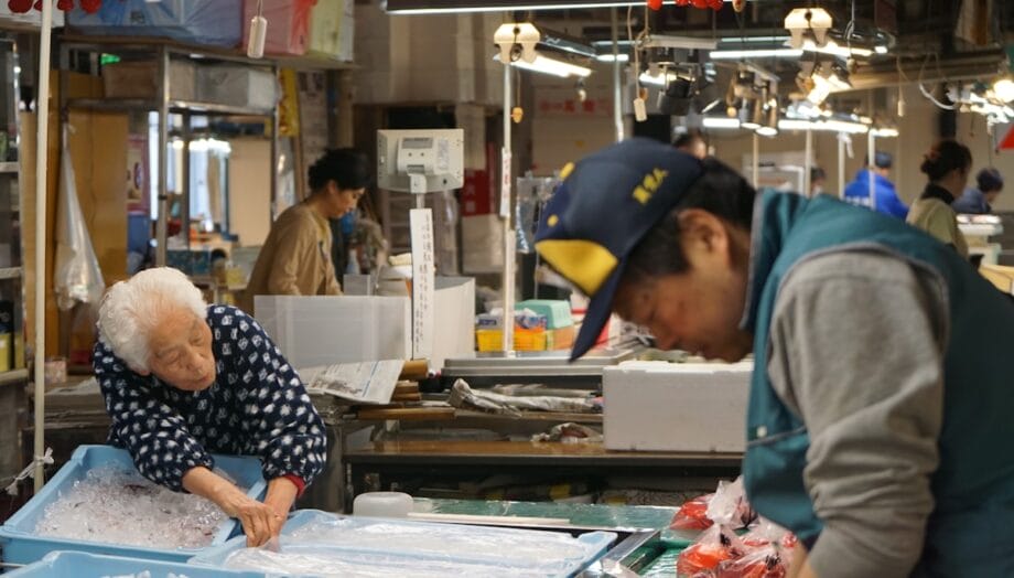 Elderly woman arranges items in a bin of ice while a worker handles goods at a busy indoor market.