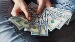 Person holding and fanning out several US 100 dollar bills while sitting with legs crossed.
