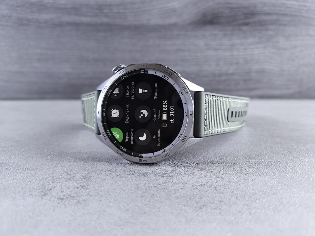 A silver smartwatch with a light gray strap displaying various health and sleep tracking icons on its screen, placed on a gray surface.