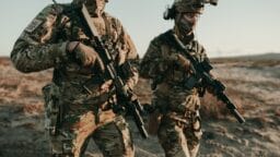 Two soldiers in camouflage uniforms and tactical gear walk outdoors, each carrying a rifle.