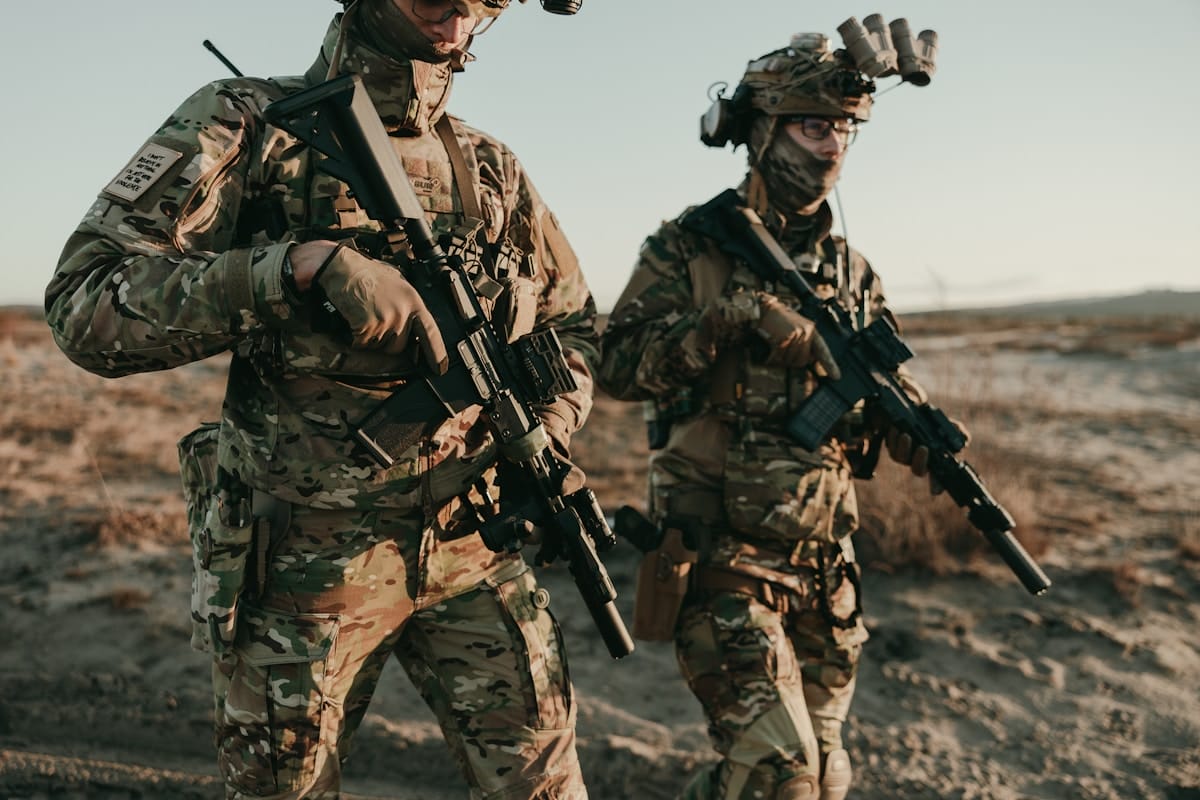 Two soldiers in camouflage uniforms and tactical gear walk outdoors, each carrying a rifle.