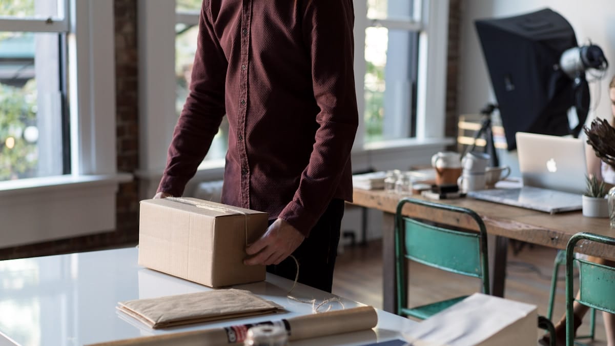 Person in a maroon shirt standing at a table, handling a cardboard box in a well-lit workspace with office supplies around.