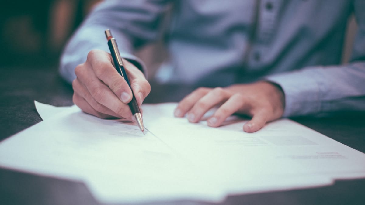 A person in a blue shirt writes on paper with a pen at a desk.