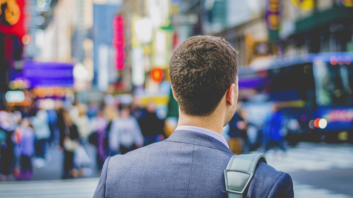 A man in a suit with a bag strap over his shoulder stands facing a busy city street filled with people and blurred lights.