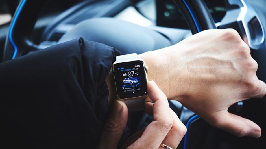 A person checks a smartwatch displaying an electric car charging status while sitting behind a car steering wheel.