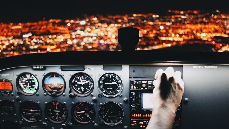 View from a small aircraft cockpit at night, with illuminated city lights below and a hand adjusting the instrument panel.