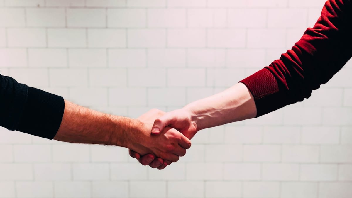 Two people are shaking hands in front of a white tiled wall.