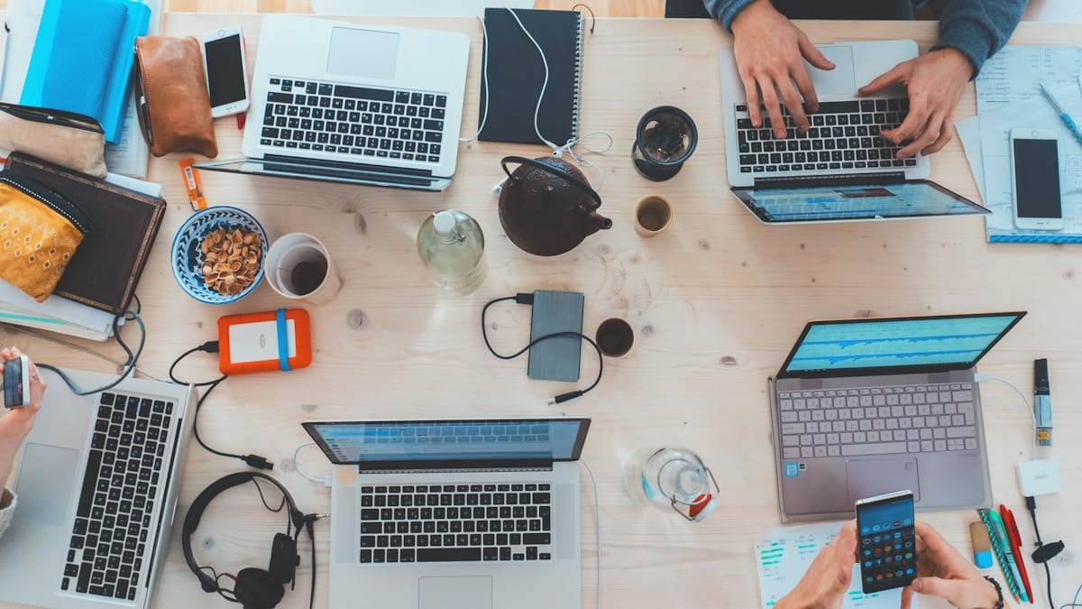 Four people working at a table with laptops, notebooks, phones, snacks, and drinks, viewed from above.