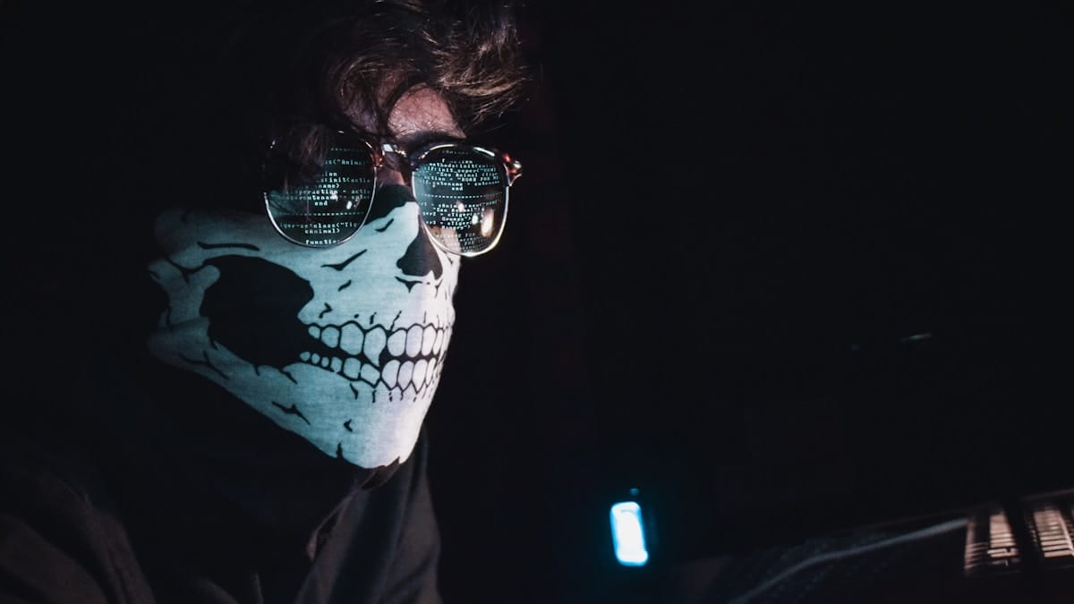 Person wearing sunglasses and a mask with a skull print, with computer code reflected in the glasses, sitting in a dark room.